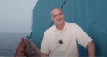 Samuele Landi, a middle-aged man smiles while sitting at the edge of his barge next to a shipping container, the ocean waves behind him.