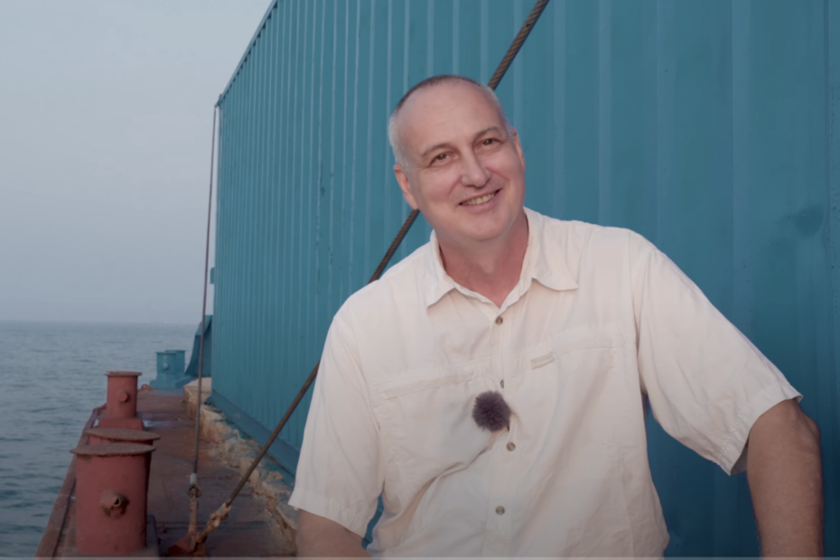 Samuele Landi, a middle-aged man smiles while sitting at the edge of his barge next to a shipping container, the ocean waves behind him.